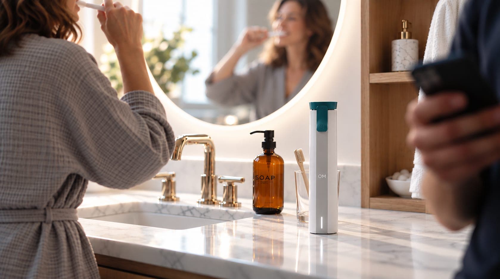 The OM reader on a sunlit marble bathroom counter, a woman brushing her teeth at the mirror beside it and someone checking a phone in the foreground.